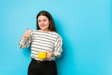 Wall Mural - Young plus size woman holding a tea mug smiling and raising thumb up