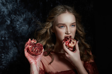 Beautiful blonde girl with pomegranate fruit in her hands. Spring portrait of a girl in a red dress breaking a pomegranate, juice flowing down her hands