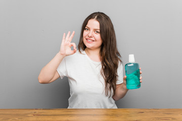 Wall Mural - Young plus size curvy woman holding a mouthwash cheerful and confident showing ok gesture.