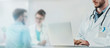 © Suteren Studio - Doctor in a white coat typing on a laptop at the hospital with nurse and patient in background. Banner crop.