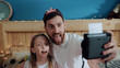 © Fractal Pictures - Funny young family man and his daughter showing funny grimace with tongues out taking selfies on polaroid camera at home.