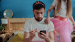 © Fractal Pictures - Portrait of bored young father using a smartphone playing with his daughter. Cheerful cute girl playing like fairy dancing around her daddy sitting with sad grimace on bed pretending to have fun.