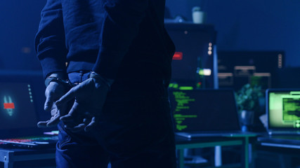 Dangerous computer hacker arrested by cyber security forces in the office at night. Close-up of hacker handcuffed against computer desk.