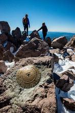 A Geological Survey Marker Marks The Summit Of Lassen Peak (Mount Lassen, 10,457 Feet) With Hikers Standing In The Background In Lassen Volcanic National Park, California.