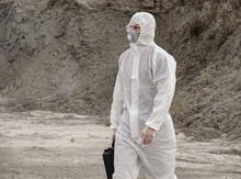 Lab Technician In A Mask And Chemical Protective Suit, Walks On Dry Ground With A Tool Box Through Toxic Smoke