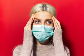 Wall Mural - Studio portrait of young blonde girl with blue eyes, having headache, holding hands on temples, wearing medical flu mask and sweater on red background.