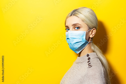 Studio profile portrait of young blonde girl wearing medical flu mask and sweater on yellow background with copy space Poster Mural XXL