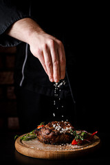 Wall Mural - The concept of cooking meat. The chef cook salt on the cooked steak on a black background, a place under the logo for the restaurant menu. food background image, copy space text