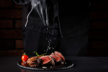 The Concept Of Cooking Meat. The Chef Cook Salt On The Cooked Steak On A Black Background, A Place Under The Logo For The Restaurant Menu. Food Background Image, Copy Space Text
