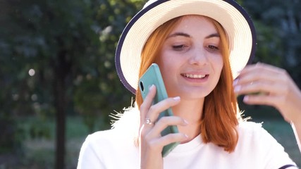 Wall Mural - Portrait of pretty positive teenage girl with red hair wearing straw hat and pink earphones talking happily on mobile phone in park.