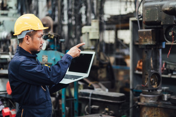 Asian Chief Engineer in the Hard Hat and working on computer laptop about mechanical piece At old Factory Equipment.