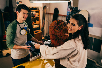 Wall Mural - worker holding terminal and smiling woman paying with credit card and holding snowboard in repair shop