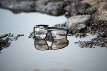 Sunset Reflected In The Lenses Of A Pair Of Glasses Placed On The Seashore Among The Lava Rocks Of Lanzarote. Spanish Island That Is Part Of The Canary Archipelago. Seascape Reflected During A Sunset