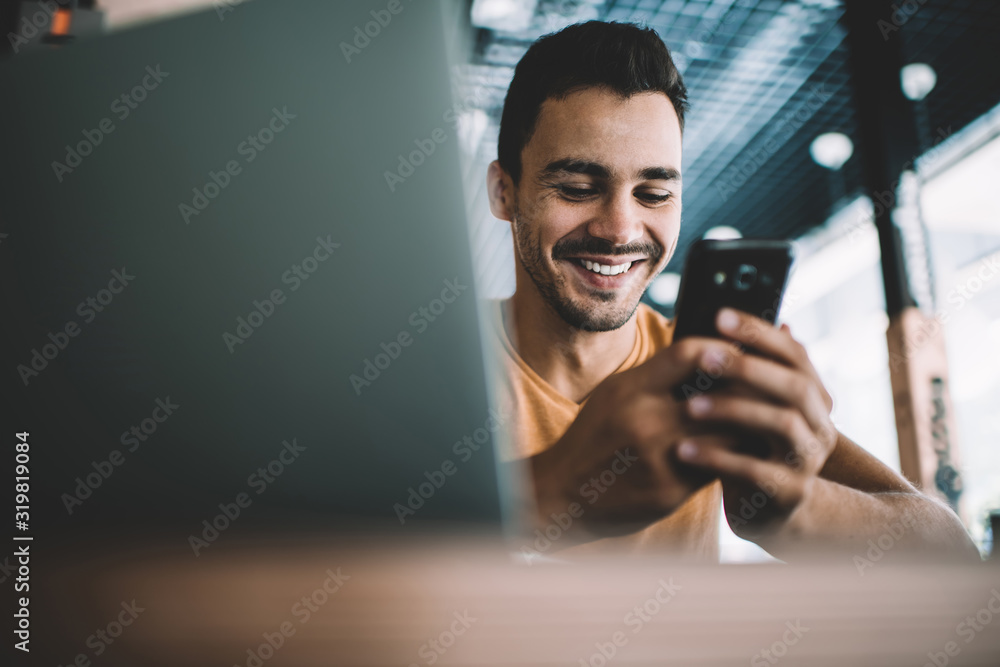  Millennial male laughing watching funny video on smartphone spending free time in cafe interior,successful man blogger satisfied with online earning getting income on banking account on cellular. - obrazy, fototapety, plakaty 