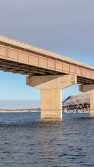 Canvas Print - Vertical frame Stringer bridge spanning over a lake with view of snowy terrain and cloudy sky