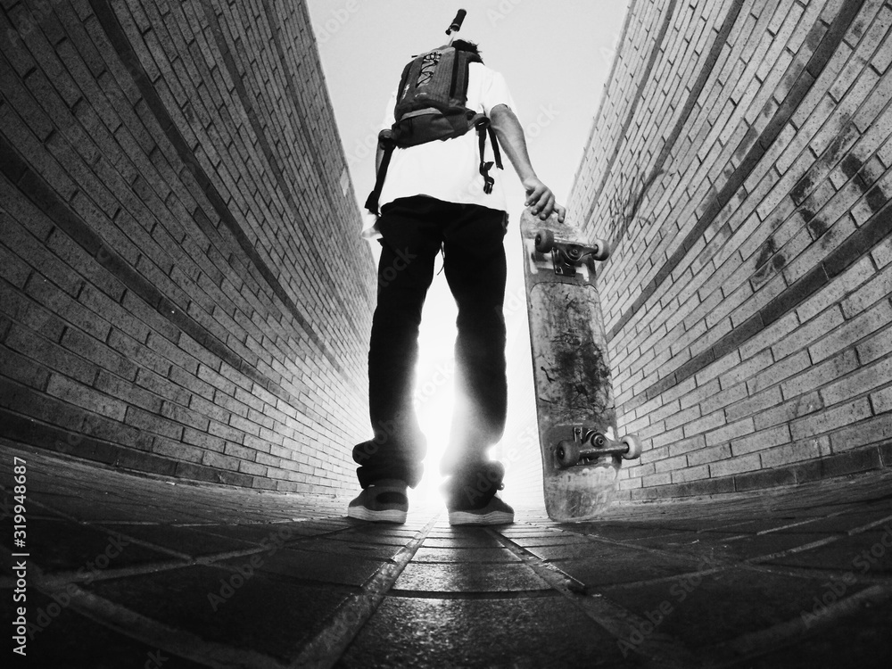Low Angle View Of Man Holding Skateboard While Standing On Footpath - obrazy, fototapety, plakaty 