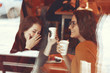 Two friends enjoying coffee together in a coffee shop viewed through glass with reflections as they sit at a table chatting and laughing