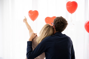 Romance moment of young Western couple, a couple sitting on the bed in bedroom with red heart shape balloons  in background. Photograph with copy space.