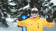 Close up portrait of bearded snowboarder or skier wearing black goggles and orange hat with snow on face standing in mountain forest and smiling and showing thumbs up sign with hands in mittens.