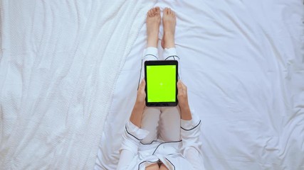female in white pajamas sits on white bed in bedroom using digital tablet with green screen