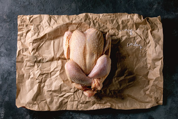 Whole raw organic uncooked farmer chicken poultry on crumpled craft paper over black concrete background. Flat lay, space