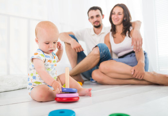  Little cute six-month-old baby collects a pyramid on the background of blurry laughing parents. Concept child care and children's health