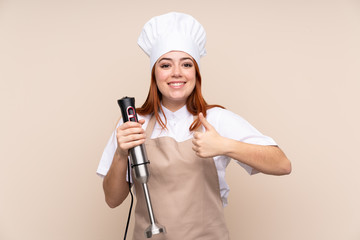 Redhead teenager girl using hand blender over isolated background with thumbs up because something good has happened