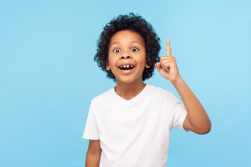 Eureka! Portrait of smart little boy with curly hair pointing finger up and looking inspired by genius thought, showing good idea sign, having clever solution in mind. studio shot blue background