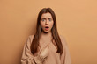 © WHstudio Leushin N - Stunned brunette European woman indicates at herself with indignant stupefied expression, shocked being chosen, frowns eyebrows, wears loose sweatshirt, models over beige wall. You blame me?