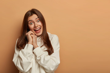 Joyful lovely woman fools around with friends, has positive gaze at camera, keeps hands near face, tilts head, wears casual white hoodie, opens mouth, poses against brown background, blank space