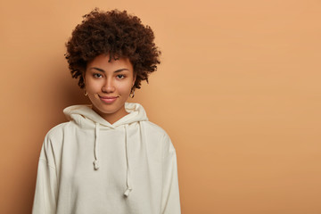 Studio shot of good looking woman has Afro hair, has direct gaze and tender smile, wears white sweatshirt, poses over beige wall with copy space for your promotion. Human facial expressions.