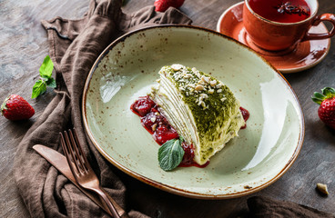 Wall Mural - Matcha green tea cake with creamy filling, nuts, strawberry jam and mint on plate over wooden background. Delicious sweets and dessert, close up