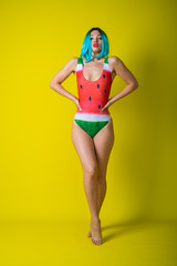 Wall Mural - Portrait of a woman in a swimsuit with a picture of a watermelon. Stylish girl in a colored short wig posing in the studio on a yellow background.