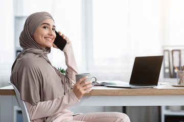 Wall Mural - Joyful arabic secretary resting at workplace with cellphone and coffee