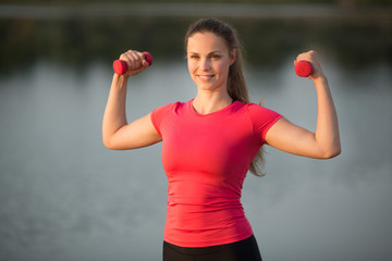 Sticker - beautiful young woman in sportswear is engaged in summer training in a park near the lake with dumbbells in her hands