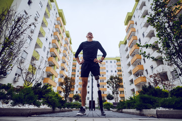 Wall Mural - Low angle view of handsome sportsman with artificial leg standing with hands on hips outdoors surrounded by buildings.