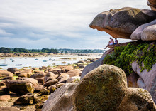 Two Young Men Sitting High Up Under A Large, Balanced Pink Granite Boulder Are Enjoying The View Over The Sainte-Anne Bay At Low Tide By A Sunny Summer Day In Tregastel, Northern Brittany, France.