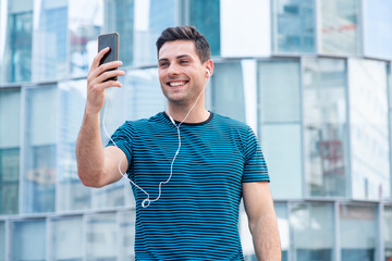 Wall Mural - smiling young man holding mobile phone taking selfie outside in city