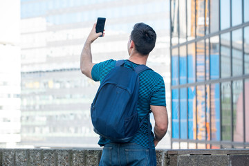 Wall Mural - behind of young man with backpack looking at city taking selfie with mobile phone