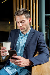 Wall Mural - young businessman sitting at city cafe with cup of coffee and looking at mobile phone