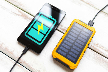 Concept of modern technologies and power bank. Smartphone with a turquoise battery icon, charged from a portable solar charger. White wooden background with stripes. Close up