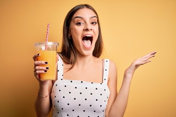 Young beautiful brunette woman drinking healthy orange juice over yellow background very happy and excited, winner expression celebrating victory screaming with big smile and raised hands