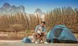 © chayathon2000 - Asian young man in blue shirt with cute puppy dog camping on the lake hill mountain view happy and enjoy life