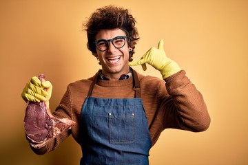 Young handsome butcher man holding meet steak standing over isolated yellow background smiling doing phone gesture with hand and fingers like talking on the telephone. Communicating concepts.