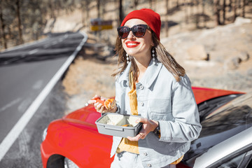 Woman having a snack with lunch box while standing on the roadside near the car, traveling on the mountains