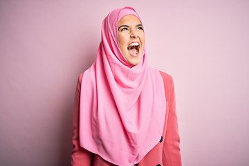 Wall Mural - young beautiful girl wearing muslim hijab standing over isolated pink background angry and mad screa