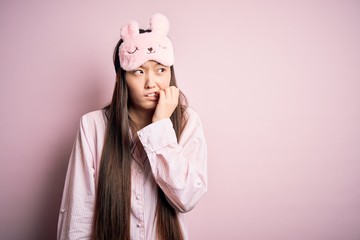 Sticker - Young asian woman wearing pajama and sleep mask over pink isolated background looking stressed and nervous with hands on mouth biting nails. Anxiety problem.