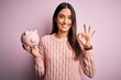 Young beautiful brunette woman holding piggy bank saving money for retirement doing ok sign with fingers, excellent symbol