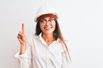 Canvas Print - Young beautiful architect woman wearing helmet and glasses over isolated white background showing and pointing up with finger number one while smiling confident and happy.