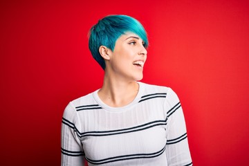 Sticker - Young beautiful woman with blue fashion hair wearing striped sweater over red background looking away to side with smile on face, natural expression. Laughing confident.